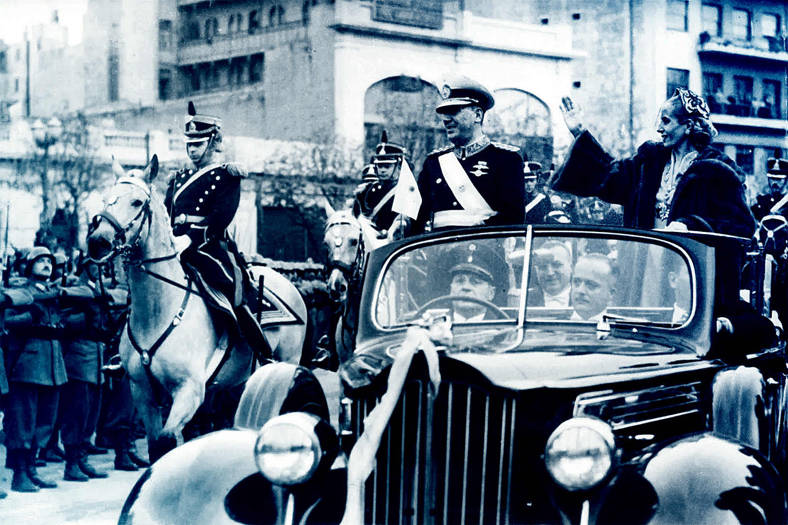 Juan and Evita Perón standing in a car waving, a soldier on horseback is beside the car