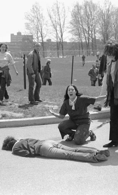 woman kneeling on ground with arms outreached near body of slain student while other students look on