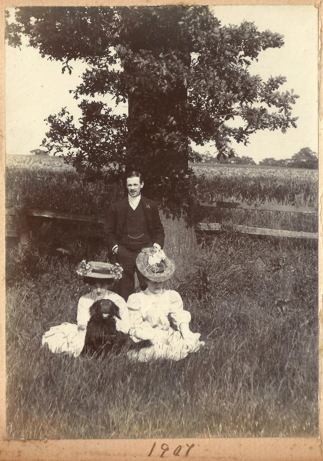 n Edwardian family poses for a family photo while on a jaunt in the countryside. Not only is the family dog included in the photograph, but they look to be having the most fun of any family member on their rural holiday. “Edwardian family group with dog, underneath the oak tree, 1907,” 1907, 