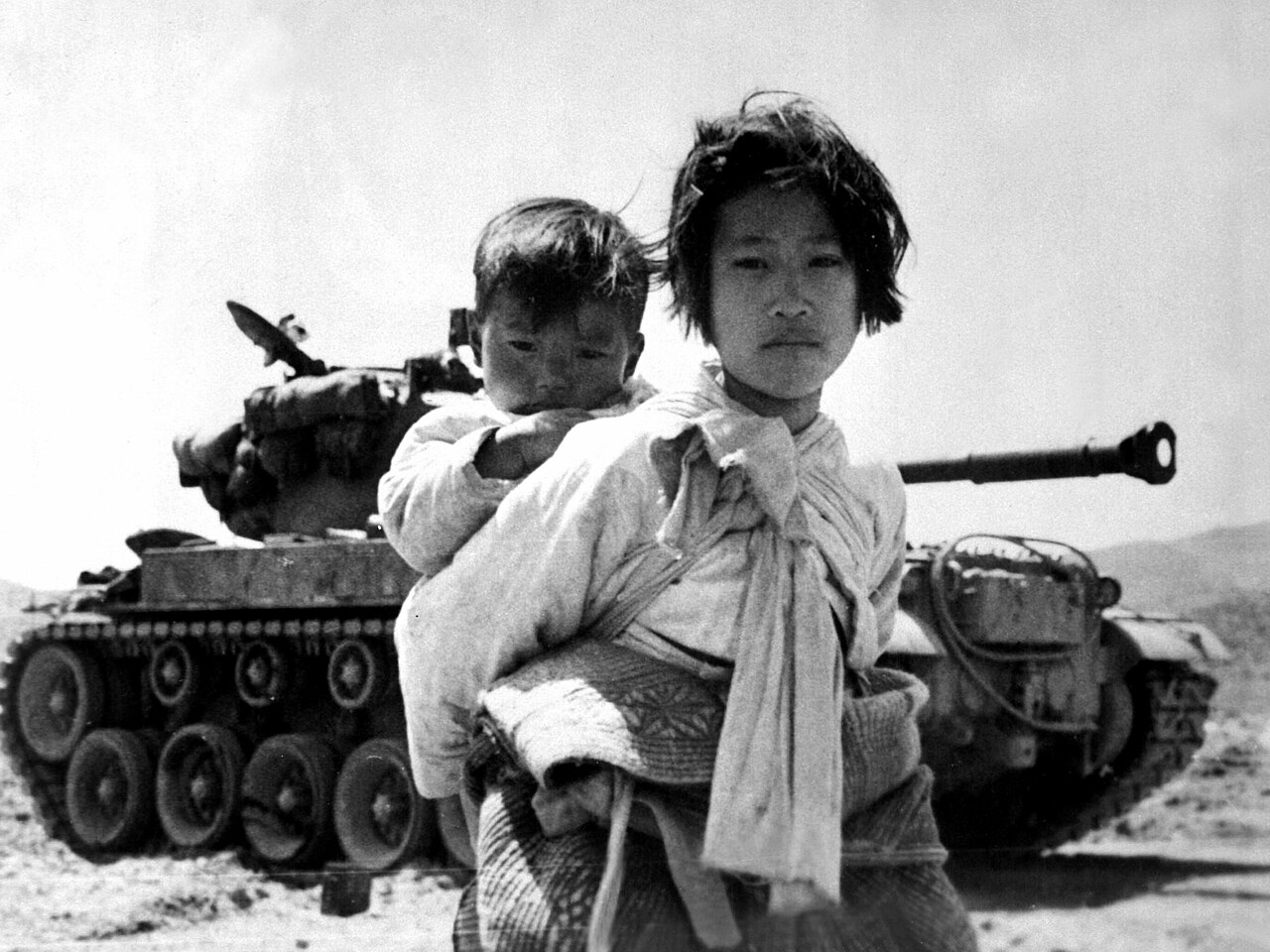 A Korean War refugee walks by a stalled M-46 tank with her brother on her back, 1951.