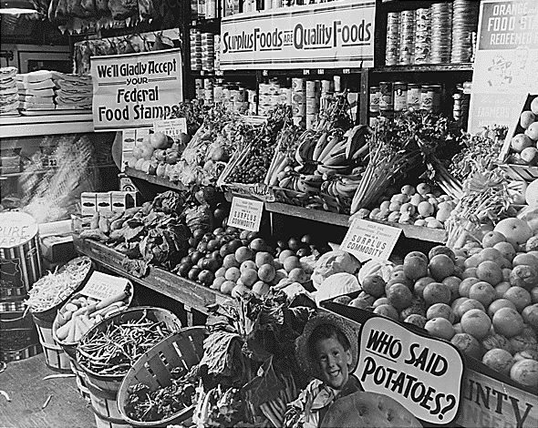 Surplus Commodities Program fresh produce display with "Surplus Foods Are Quality Foods" and "We Gladly Accept Your Federal Food Stamps" signs.