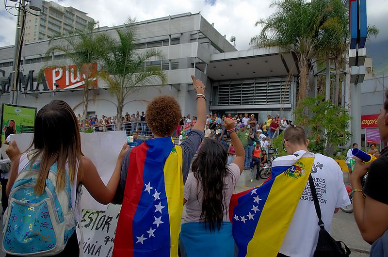 Protestors demonstrating while people seeking to buy basic goods in short supply line up outside a store in 2015.