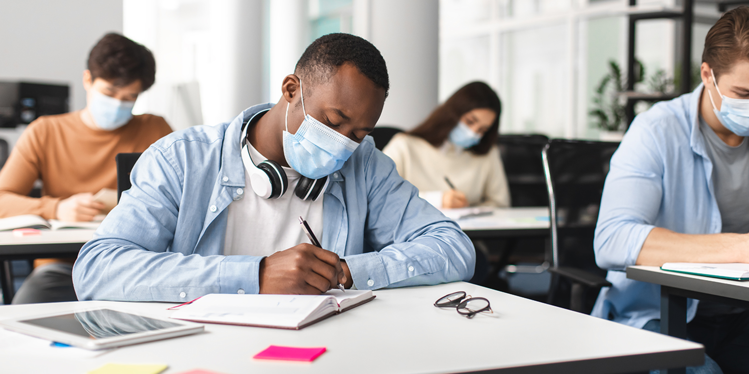 students wearing masks sitting at desks in a classroom writing