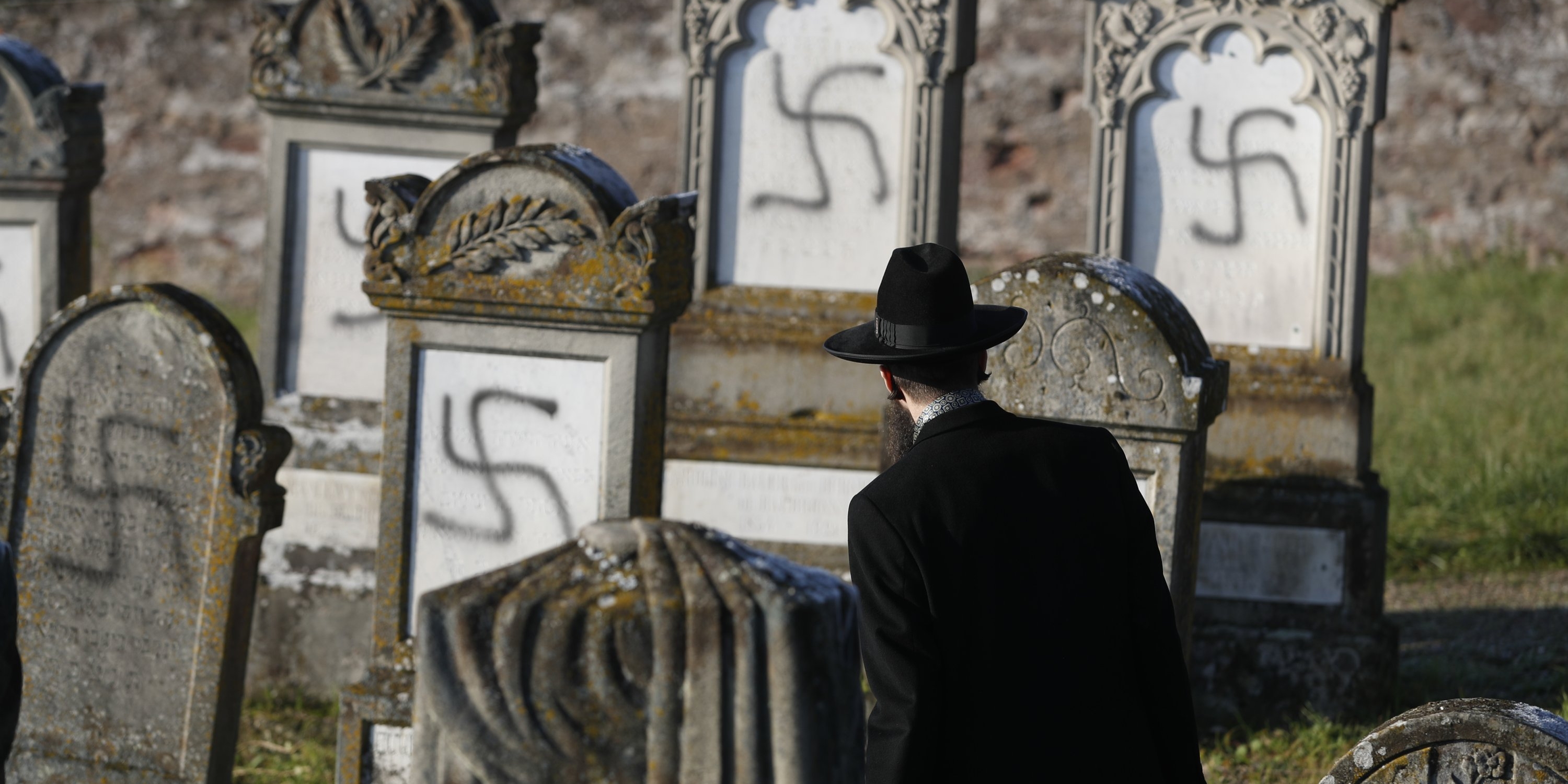 A Jewish man looking at gravestones that have been spray painted with swastikas