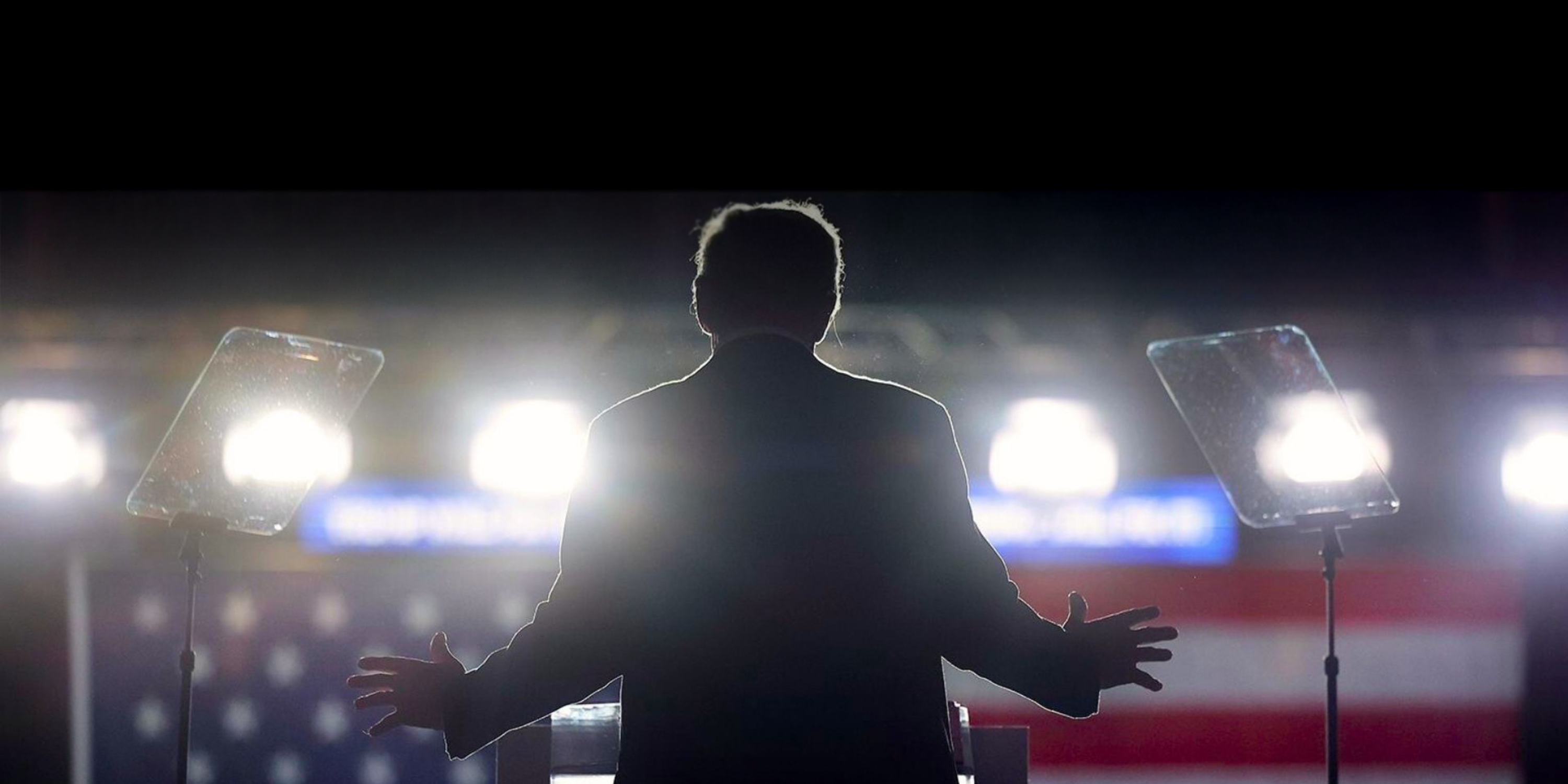 the back of Donald Trump standing in front of a U.S. flag and bright lights