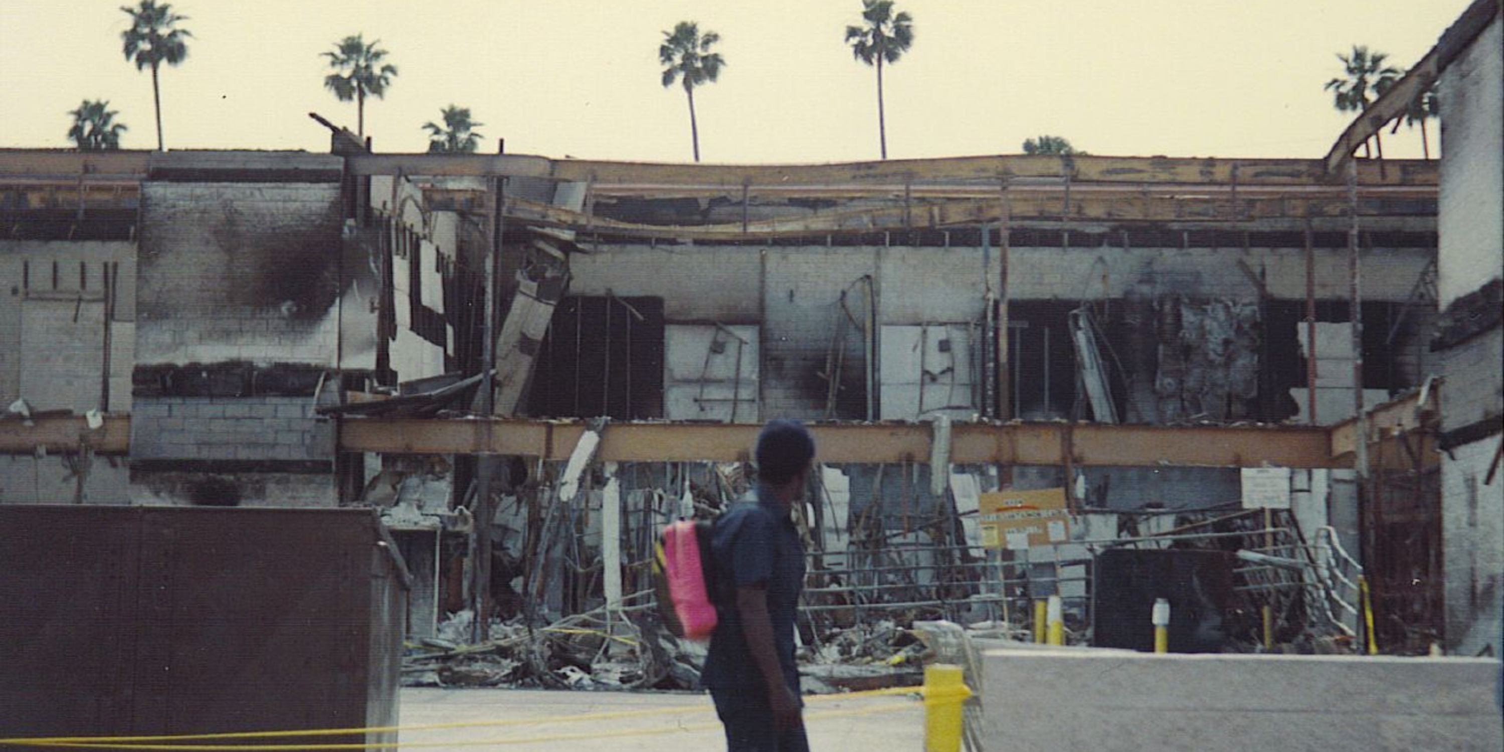 a man walking in front of burned out buildings with palm trees behind the buildings