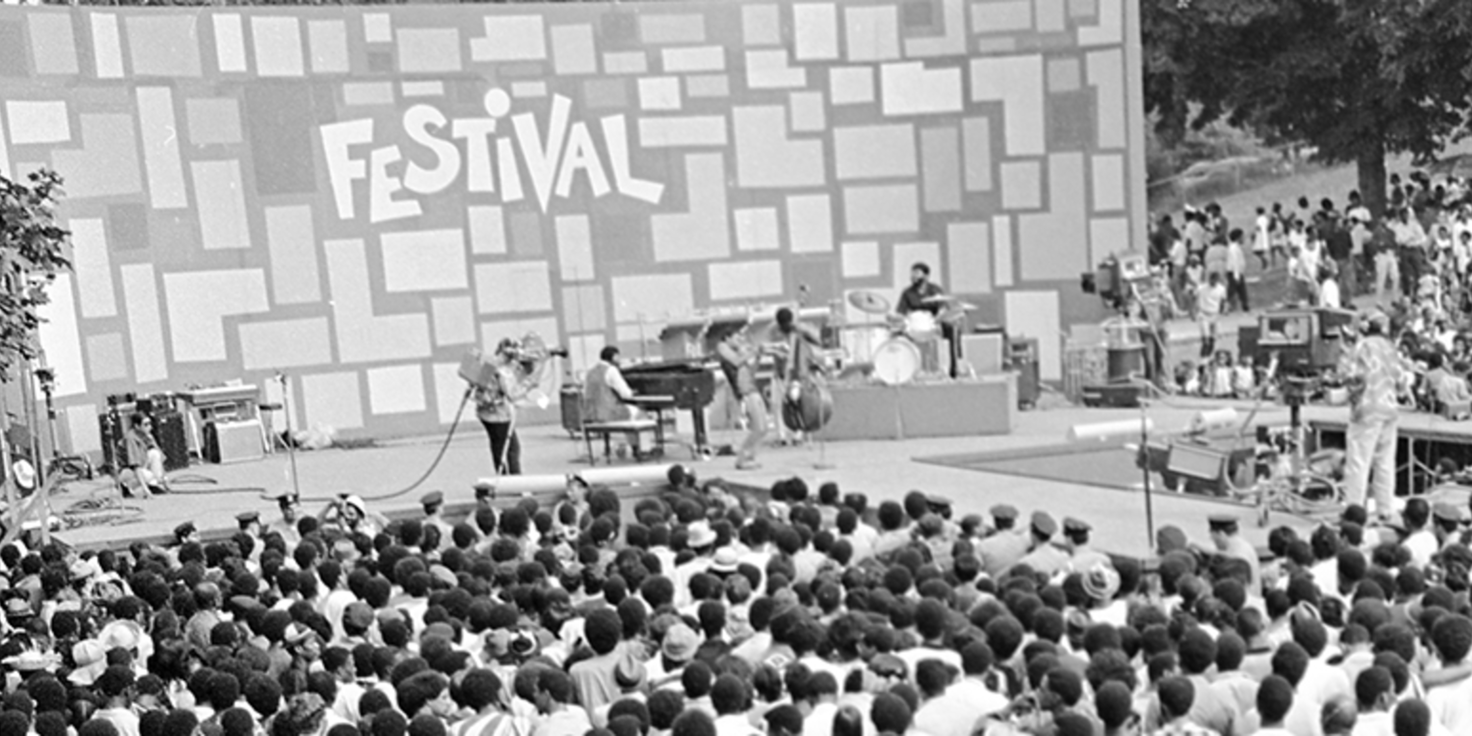 Black and white panorama photo of a stage with musical performers surrounded by a crowd