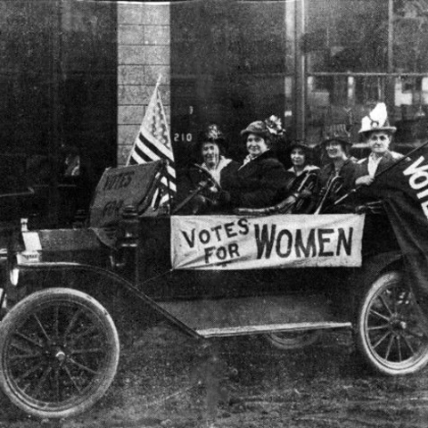 suffragettes in a car with U.S. flag and two signs that read Votes for Women