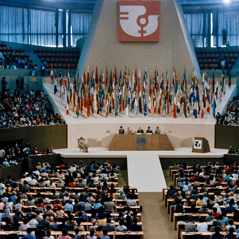 Scene from the 1975 Conference on Women. Interior of a large auditorium. Panel of people are sitting on a stage. The conference logo of a women's symbol and bird are above the stage.