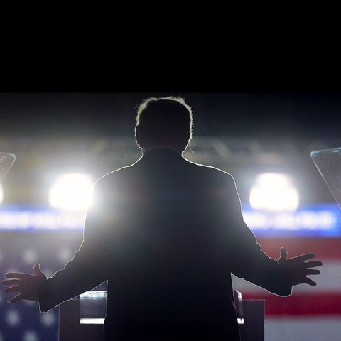the back of Donald Trump standing in front of a U.S. flag and bright lights
