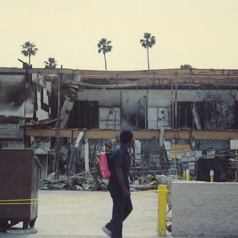 a man walking in front of burned out buildings with palm trees behind the buildings