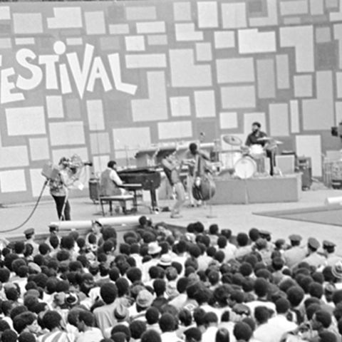 Black and white panorama photo of a stage with musical performers surrounded by a crowd