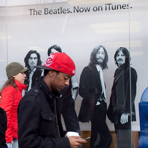 A young man stays connected on his cell phone as he strolls by a store advertising the Beatles' music on Apple's online store.