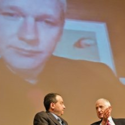 Julian Assange on screen overlooking two men talking