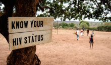 Background: children playing; foreground sign: "Know Your HIV Status"